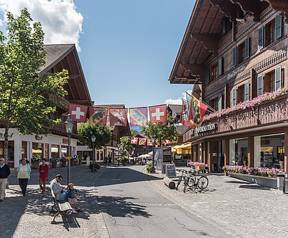 Promenade mit blumengeschmückten Chalets, Fussgängern, einem Mann und einem Kind auf einer Sitzbank, Bäume und Strassenlaternen, in der Mitte hängen Fahnen von der Schweiz, dem Kanton Bern und der Gemeinde Saanen.