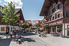 Promenade où se trouvent des chalets fleuris, des piétons, un homme et un enfant sur un banc, des arbres et des lampadaires. Au centre, la promenade est pavoisée des drapeaux de la Suisse, du canton de Berne et de la commune de Saanen.
