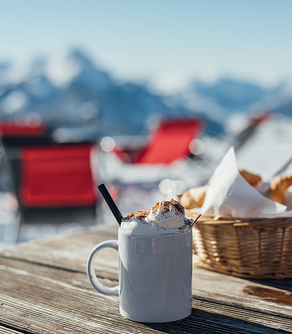 On a table is a hot chocolate with whipped cream and a basket of chips. Behind it you can see a blurry snow-covered mountain panorama.