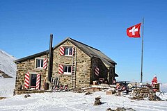 Stone SAC hut in the snow, with a Swiss flag, sun terrace and mountain panorama under a clear blue sky.