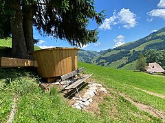 Ein überdimensionales Fondue-Caquelon steht auf einer Wiese in Abländschen. Im Hintergrund sind Berge, ein Bauernhaus und eine sonnige alpine Landschaft zu sehen.