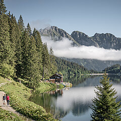Zwei Wandrer:innen auf einem Fussweg entlang eines Bergsees, im Hintergrund Berge mit einer Nebelbank davor.