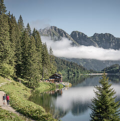Zwei Wandrer:innen auf einem Fussweg entlang eines Bergsees, im Hintergrund Berge mit einer Nebelbank davor.