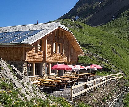 Ein gemütliches Bergrestaurant mit Holzfassade und Sonnenschirmen. Die grüne Landschaft und die Berge im Hintergrund schaffen eine einladende Atmosphäre.