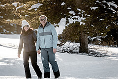  Couple walking hand in hand along a winter hiking trail, against a snowy mountain backdrop. 