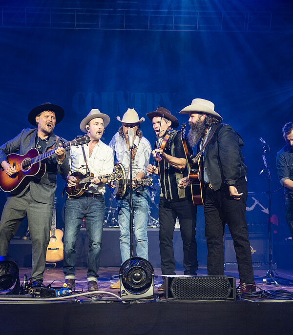 Country-Band mit Gitarre, Geige und Kontrabass auf der Bühne der Country Night Gstaad vor blau beleuchteter Konzertkulisse.